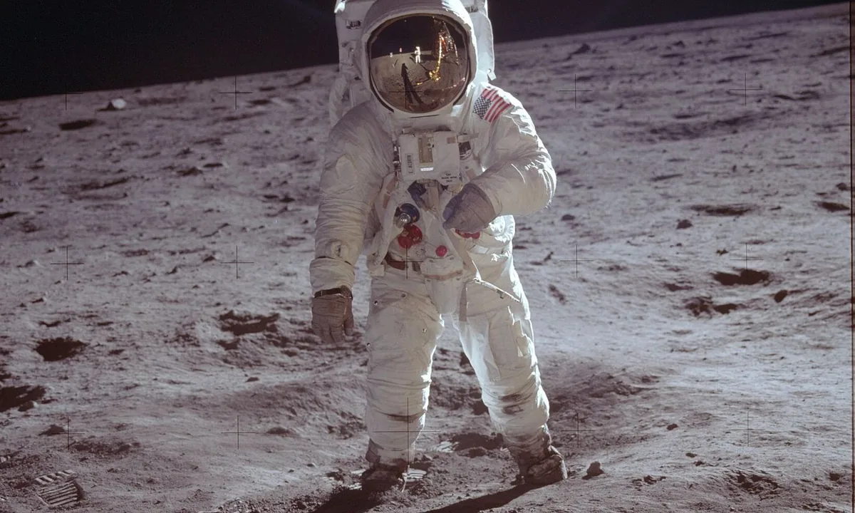 Colour photograph of an astronaut in a white spacesuit standing on grey lunar soil, with a photographer and the lunar module reflected in his gold visor.