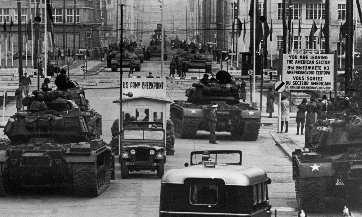 Black-and-white photo of American tanks and a jeep lined up on a Berlin street beside a "US Army Checkpoint" hut and a sector sign.