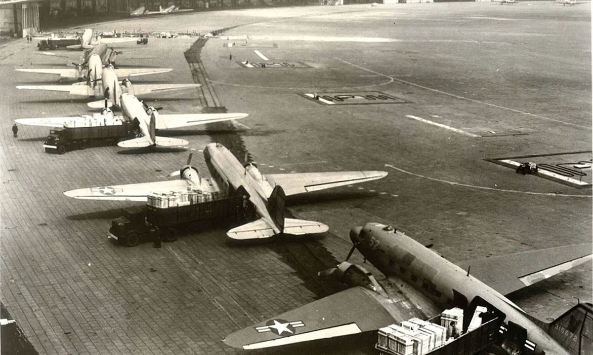 Black-and-white aerial photo of a row of twin-engine cargo planes parked on an apron with trucks loading crates beside them.