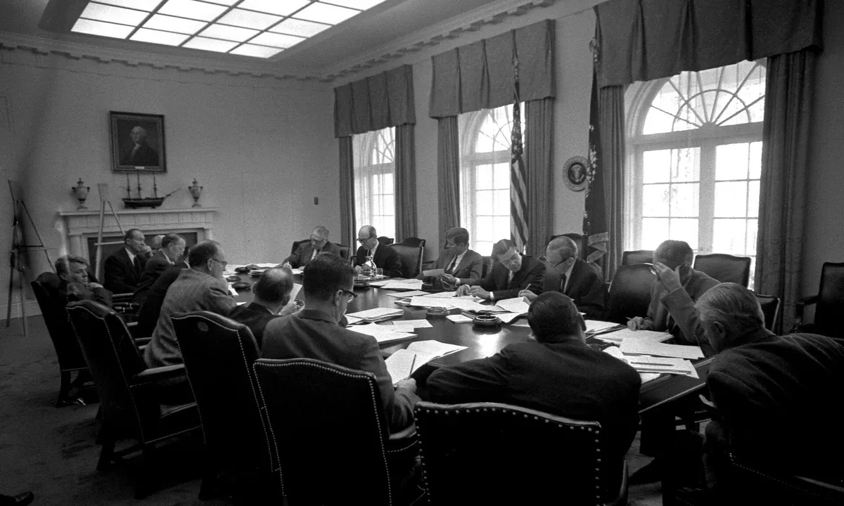 Black-and-white photo of men in suits seated around a long table covered with papers in a room with tall windows and flags.