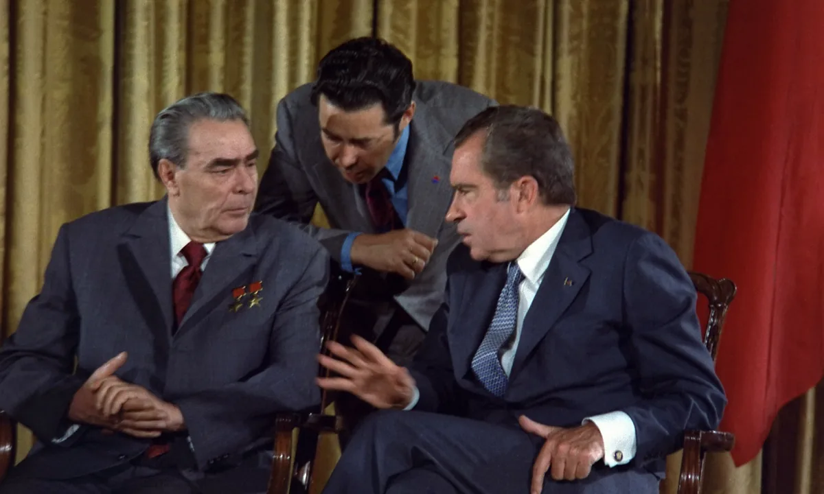 Colour photo of Brezhnev and Nixon seated in conversation while an interpreter leans in between them.