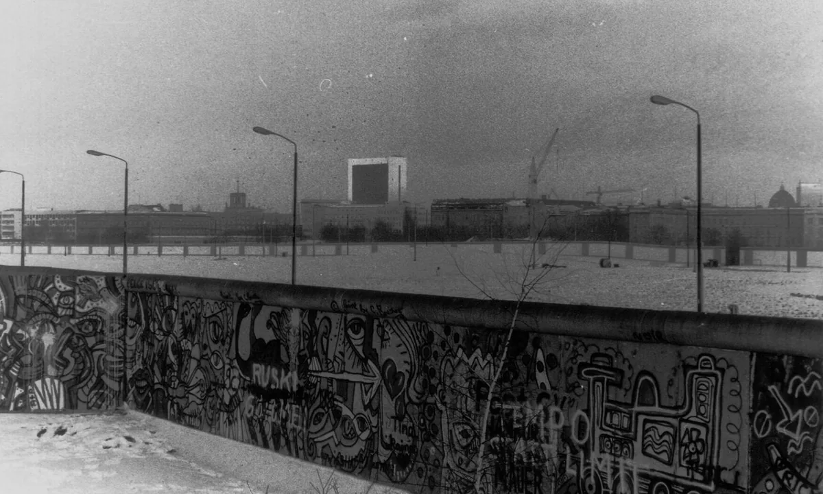 Black-and-white photo of a graffiti-covered concrete wall with an empty strip of land, lamp posts and buildings behind it.