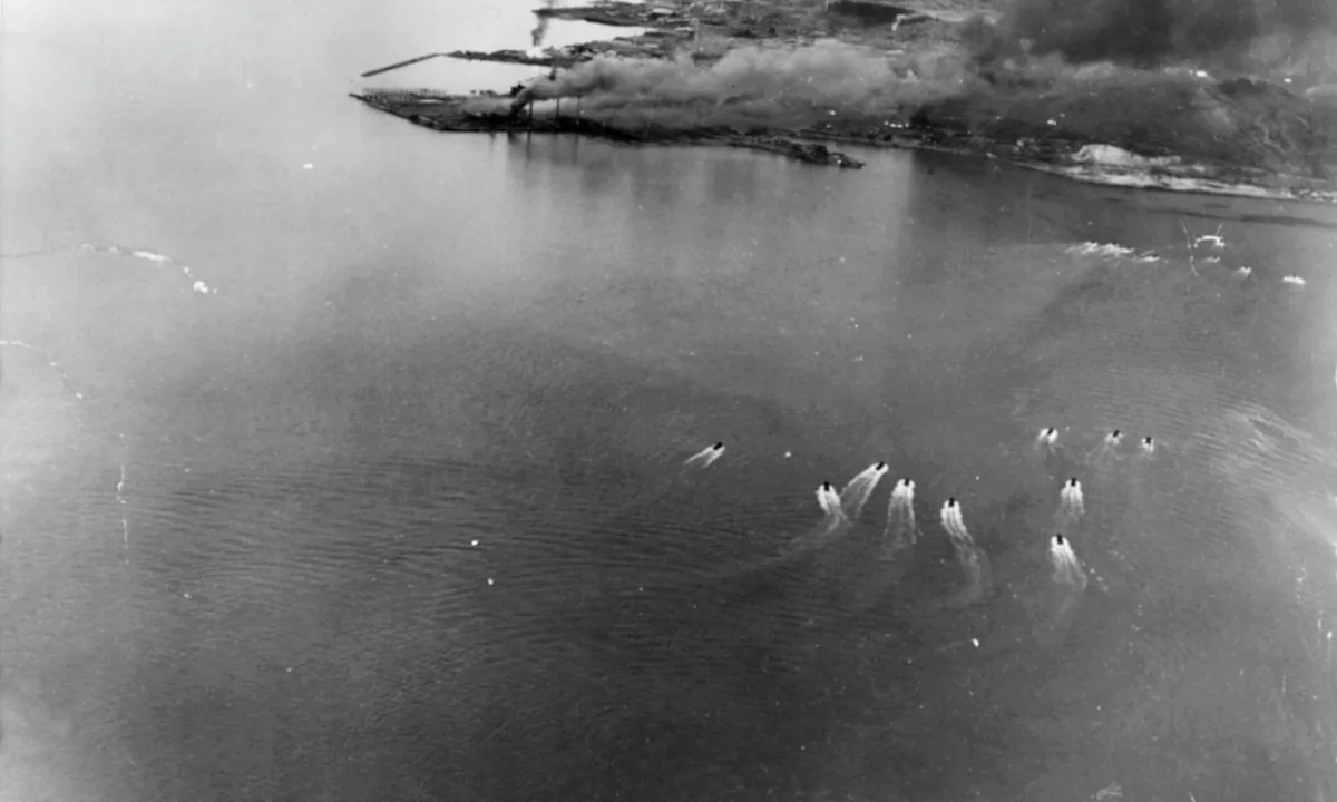 Black-and-white aerial photo of small boats leaving white wakes across a bay towards a burning harbour.