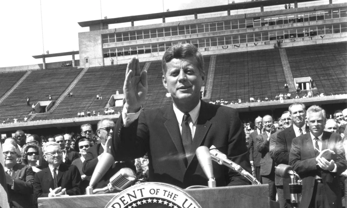 Black-and-white photograph of a man in a dark suit speaking at a podium in a stadium, with a crowd behind him.