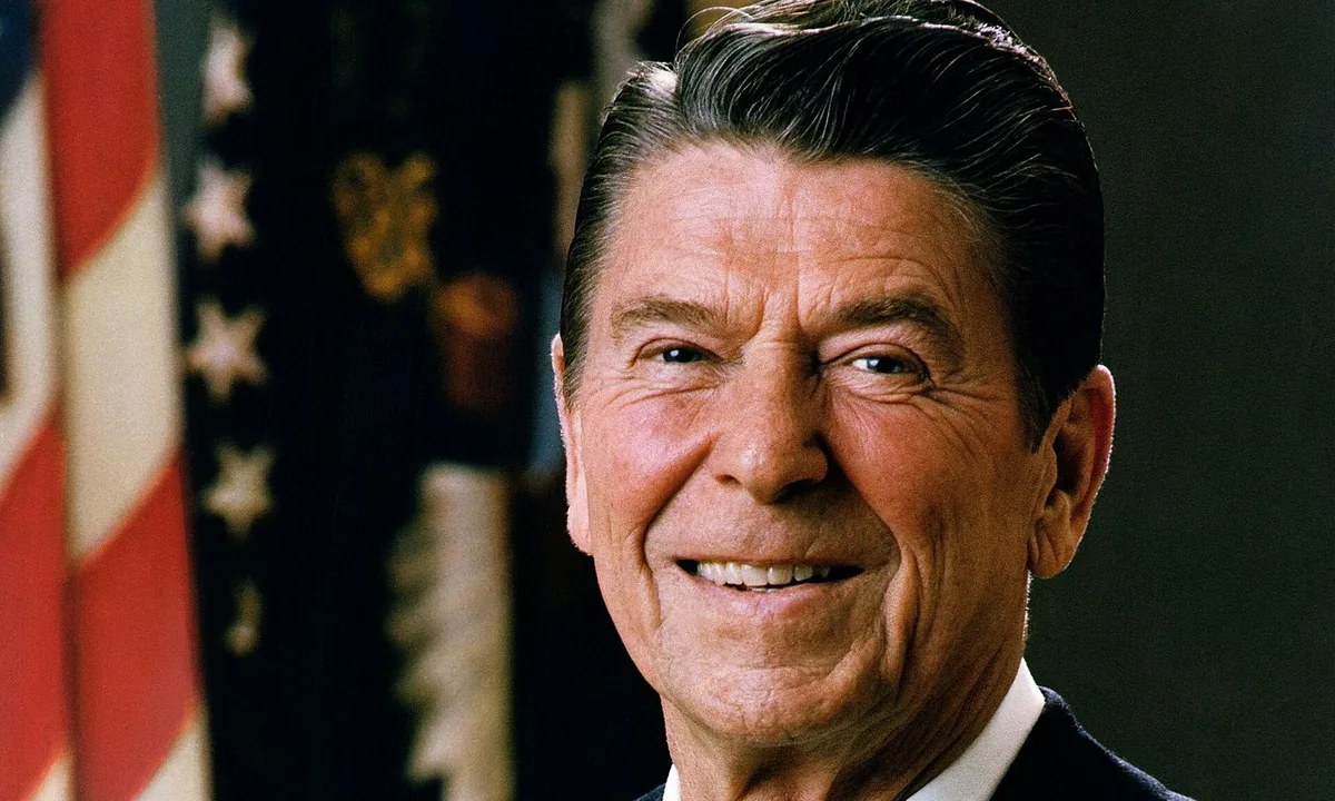 Colour portrait of Reagan, smiling, in a dark suit in front of the US flag.