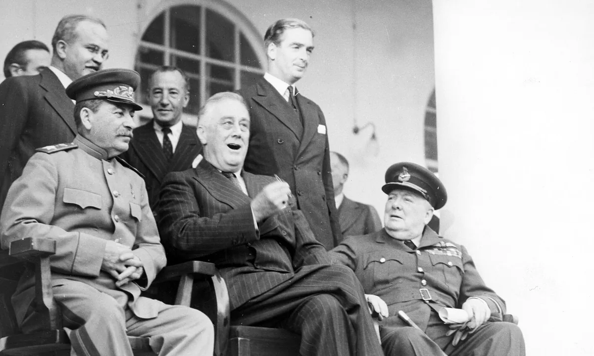 Black-and-white photo of three seated leaders: Stalin in a military cap and uniform on the left, Roosevelt laughing in the middle and Churchill in an air force uniform on the right, with aides standing behind.