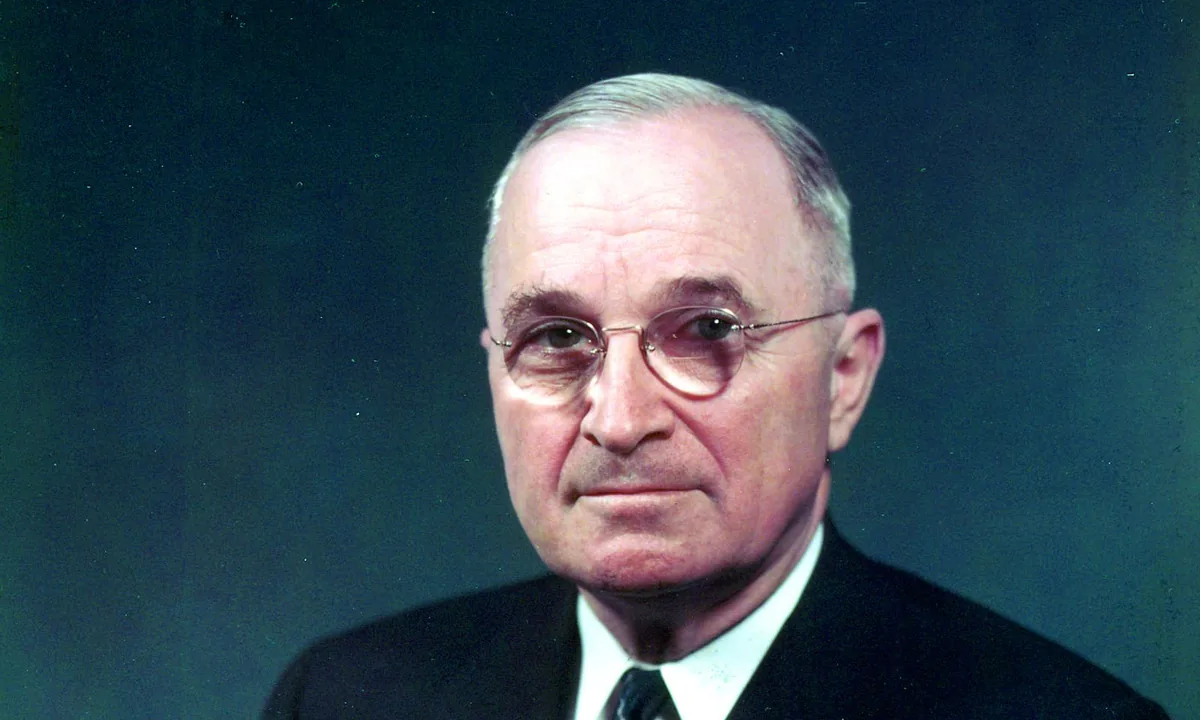Colour portrait of Truman in round glasses and a dark suit against a teal background.
