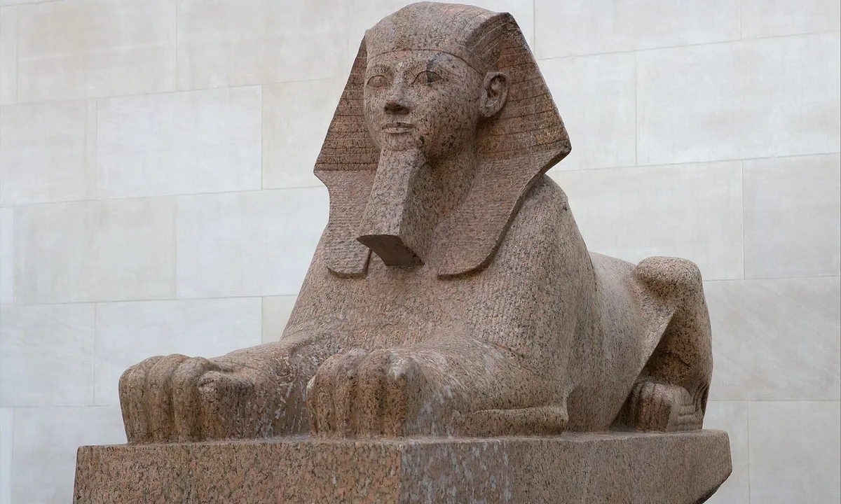 Red granite sphinx with a lion's body and a royal head wearing a striped headcloth and a long beard.
