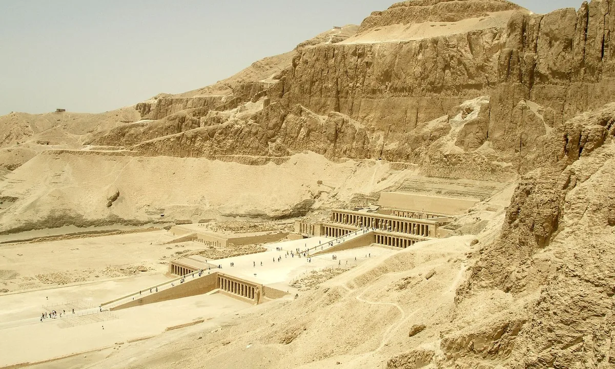 A long, pale, three-level temple with rows of columns and ramps, built against towering desert cliffs.