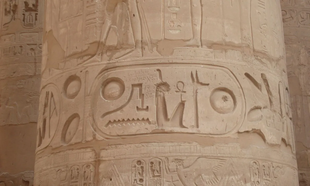 Close view of a huge sandstone column carved with large hieroglyphs, including an oval ring enclosing a sun disk, a seated goddess and other signs.