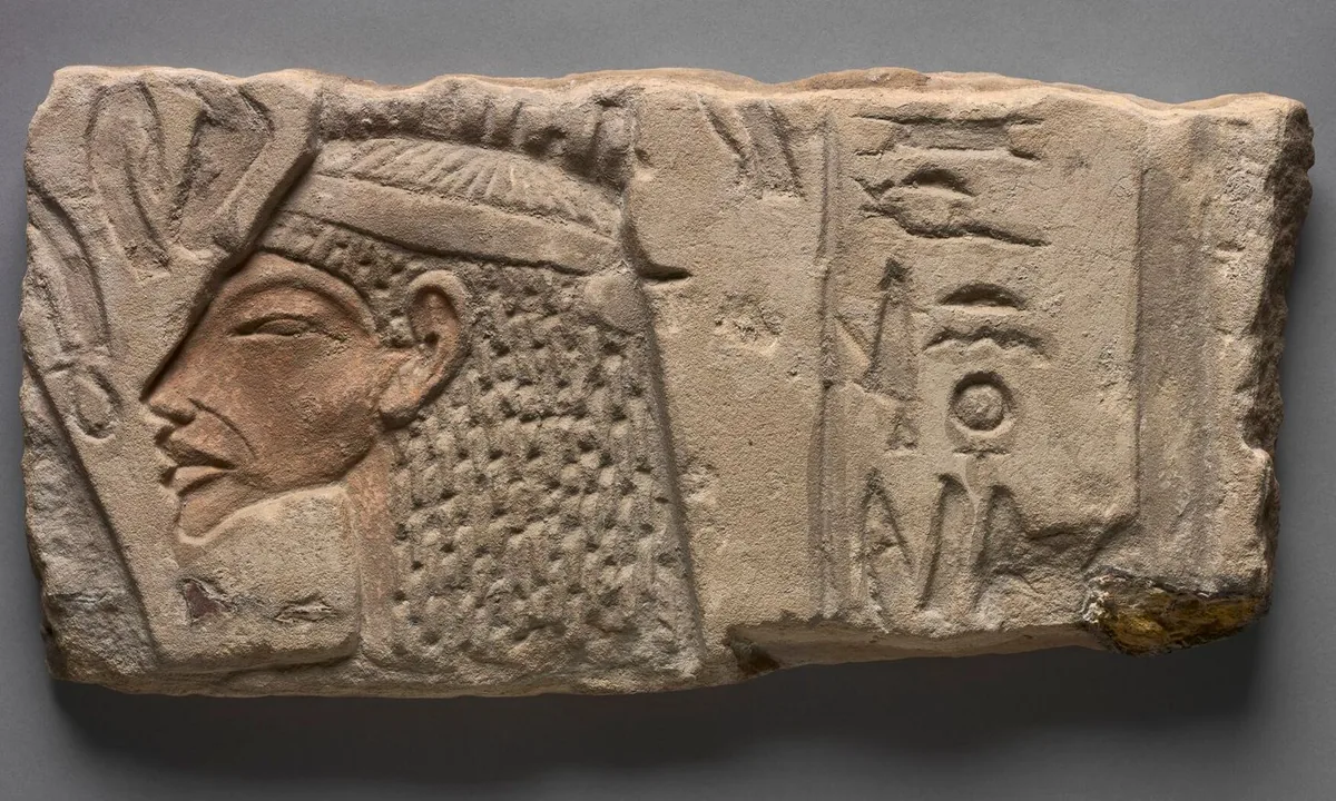 Carved sandstone block with the profile head of a queen in a close-fitting crown, beside a column of hieroglyphs.