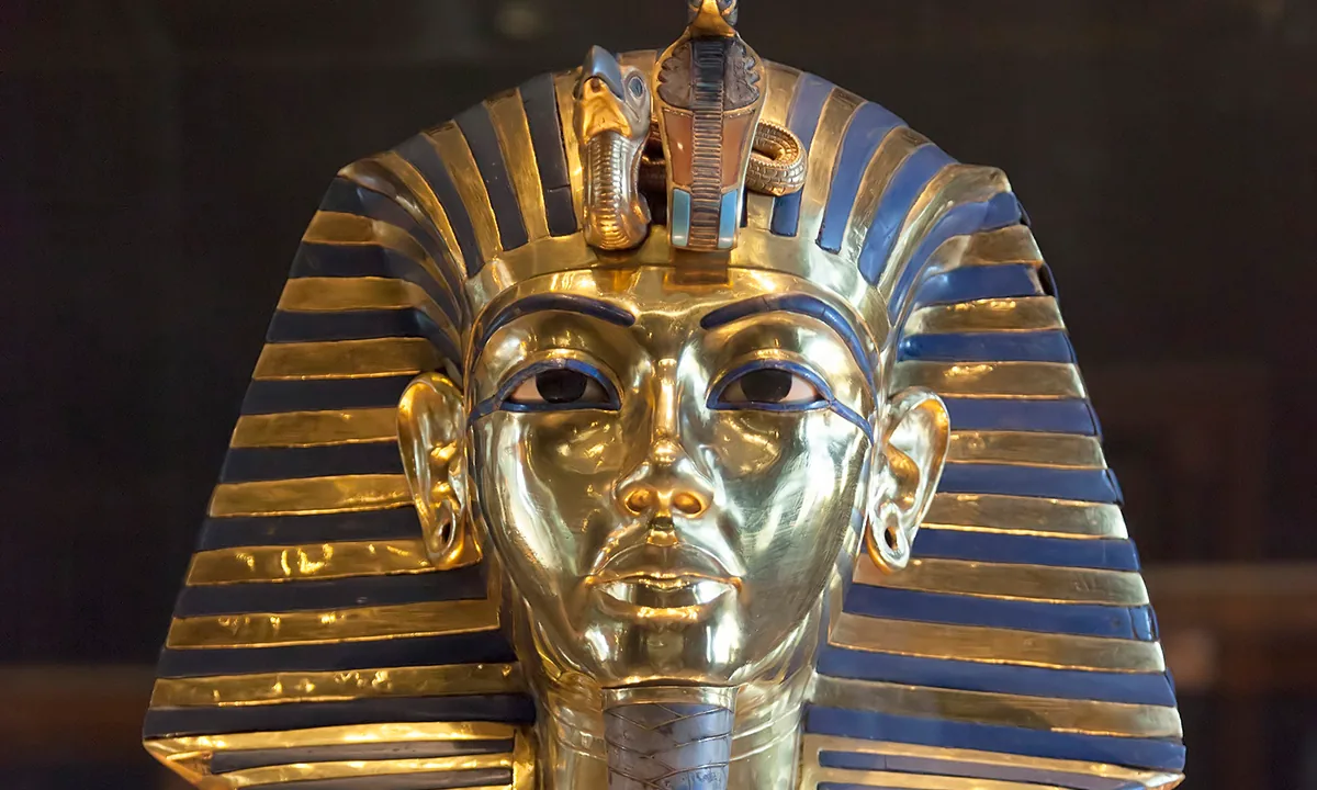 Gold mask of a young king wearing a striped blue-and-gold headcloth, with a cobra and vulture on the brow and a long braided beard.