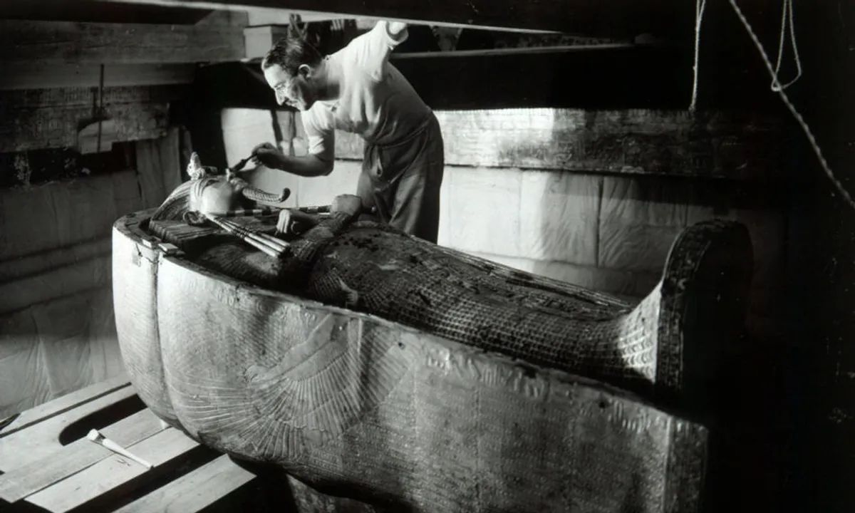 Black-and-white photograph of a man kneeling beside a gleaming gold coffin shaped like a human figure, holding a small tool.