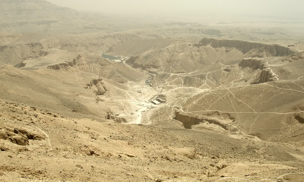 A hazy view down into a dry, branching desert valley ringed by limestone cliffs, with paths and small structures on the valley floor.