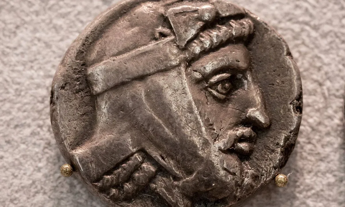Close-up of an ancient silver coin with the profile of a bearded man wearing a soft Persian cap tied with a band.