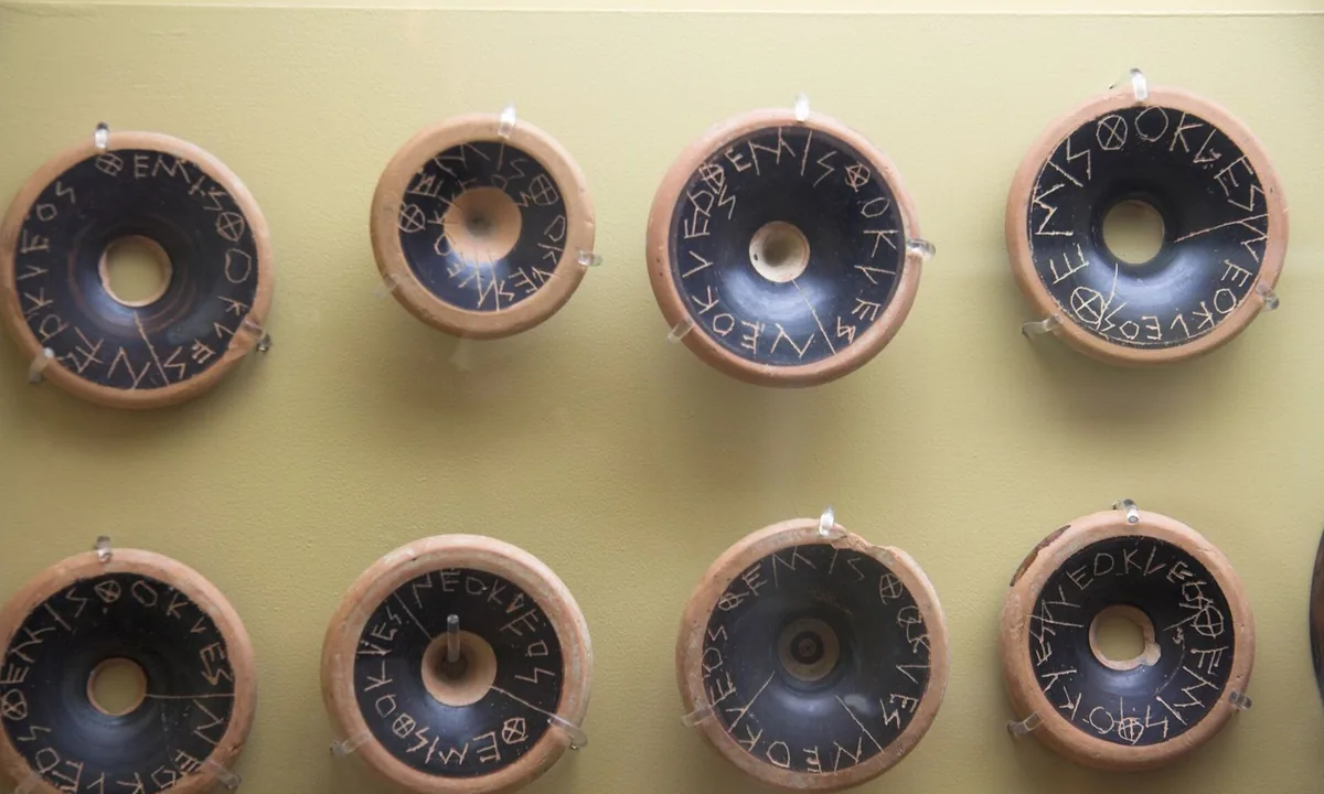 Rows of round black-glazed pottery bases mounted on a yellow wall, each scratched with Greek letters spelling the name Themistocles.