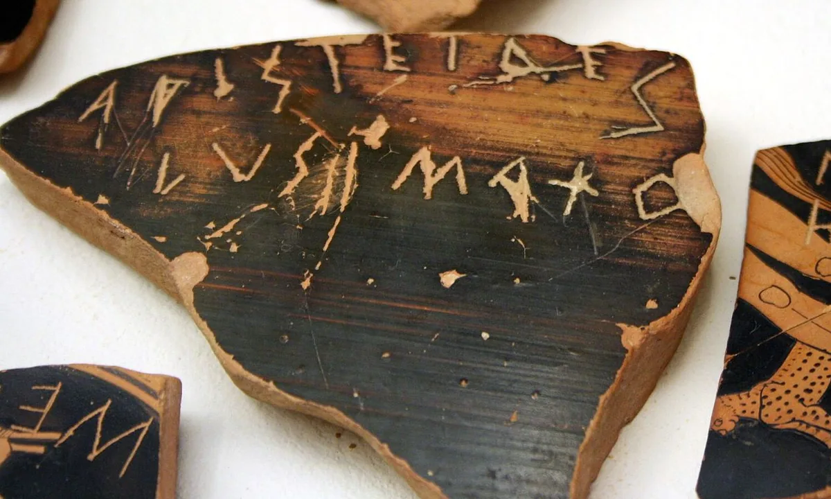 A broken piece of black-glazed pottery with Greek letters scratched across it, lying among other inscribed sherds.