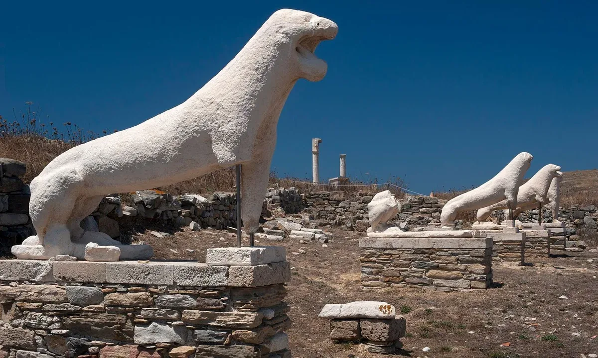 Weathered white lion statues in a row on stone bases among ruins under a deep blue sky.