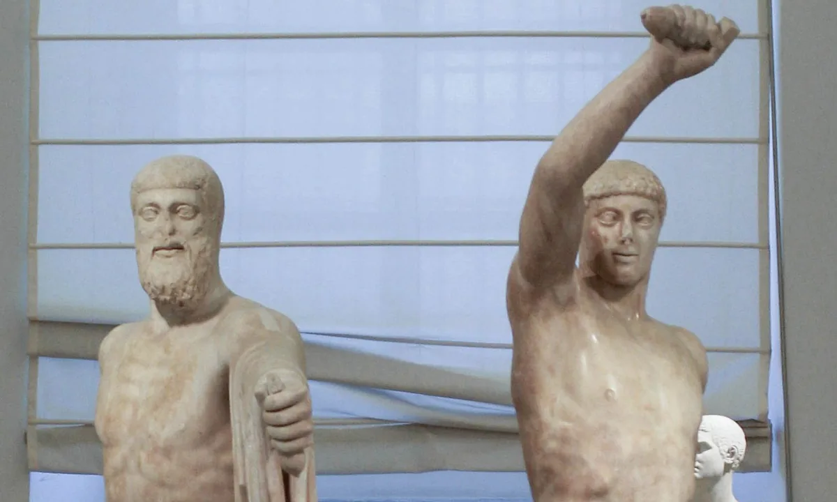 Two nude marble statues of men striding forward side by side, the younger raising one arm high above his head, the older and bearded one with a cloak over his arm.