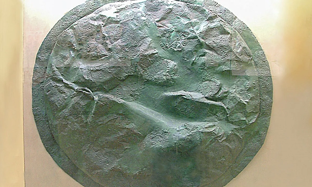 A large round bronze shield, dented and green with corrosion, displayed against a pale wall.