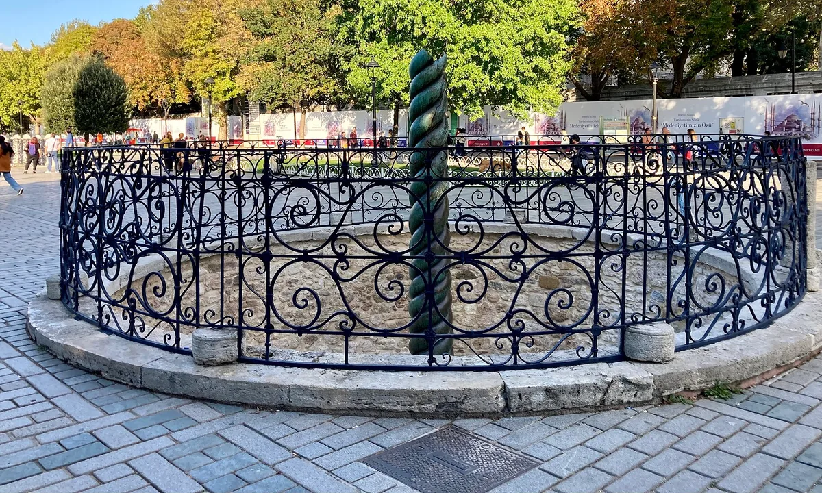 A twisted dark bronze column rising from a sunken circular enclosure surrounded by an iron railing in a paved square.