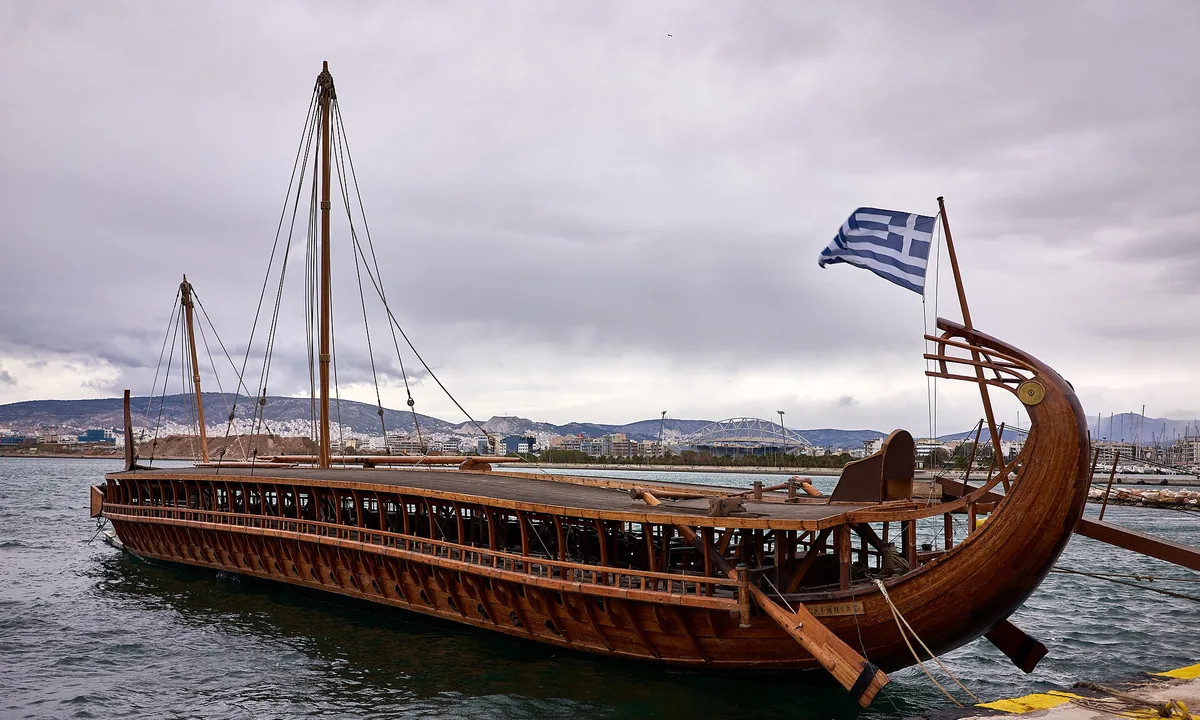 A long wooden warship with a curved stern, a painted eye on the bow and two masts, moored at a quay under a cloudy sky.