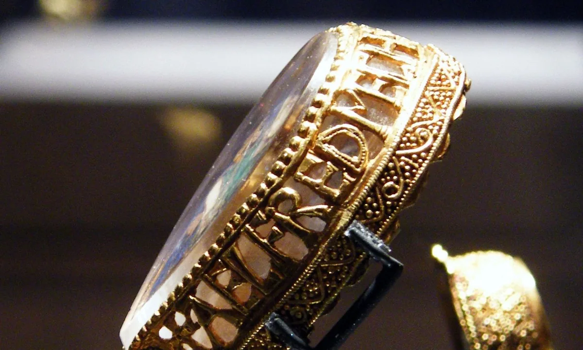 Side view of a small gold jewel: openwork gold letters run around the edge of a clear rock-crystal front, ending in a gold animal-head socket.