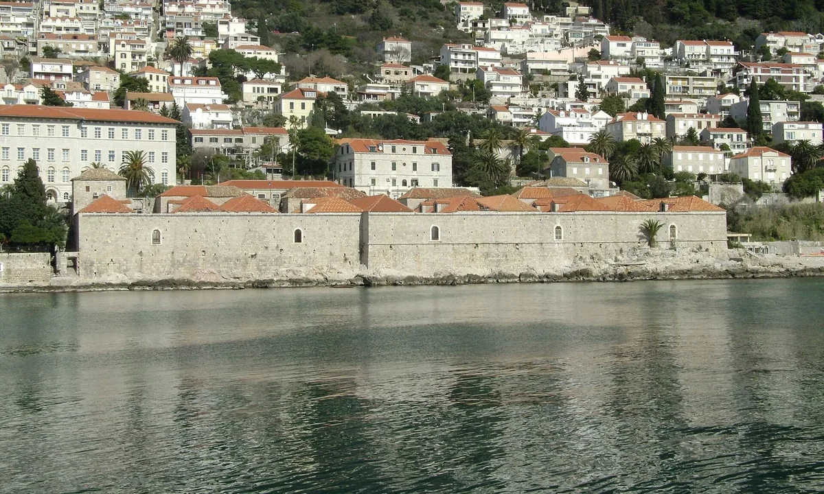 Row of long stone buildings with tiled roofs and arched doorways along a rocky seafront outside old city walls.