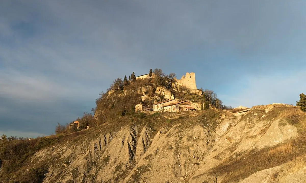 Ruined stone walls on top of a steep white rocky outcrop above green wooded hills.