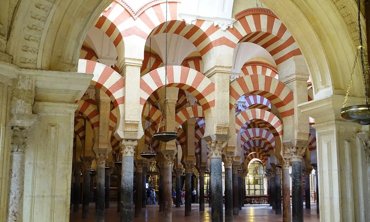 Interior of a mosque hall with rows of dark columns carrying two levels of arches built of alternating red and white stripes.