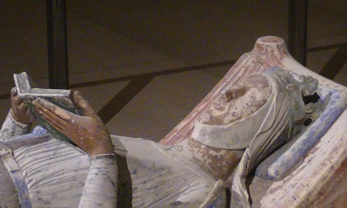 Close view of a painted stone effigy of a crowned woman in a white wimple, lying with an open book held up in both hands.