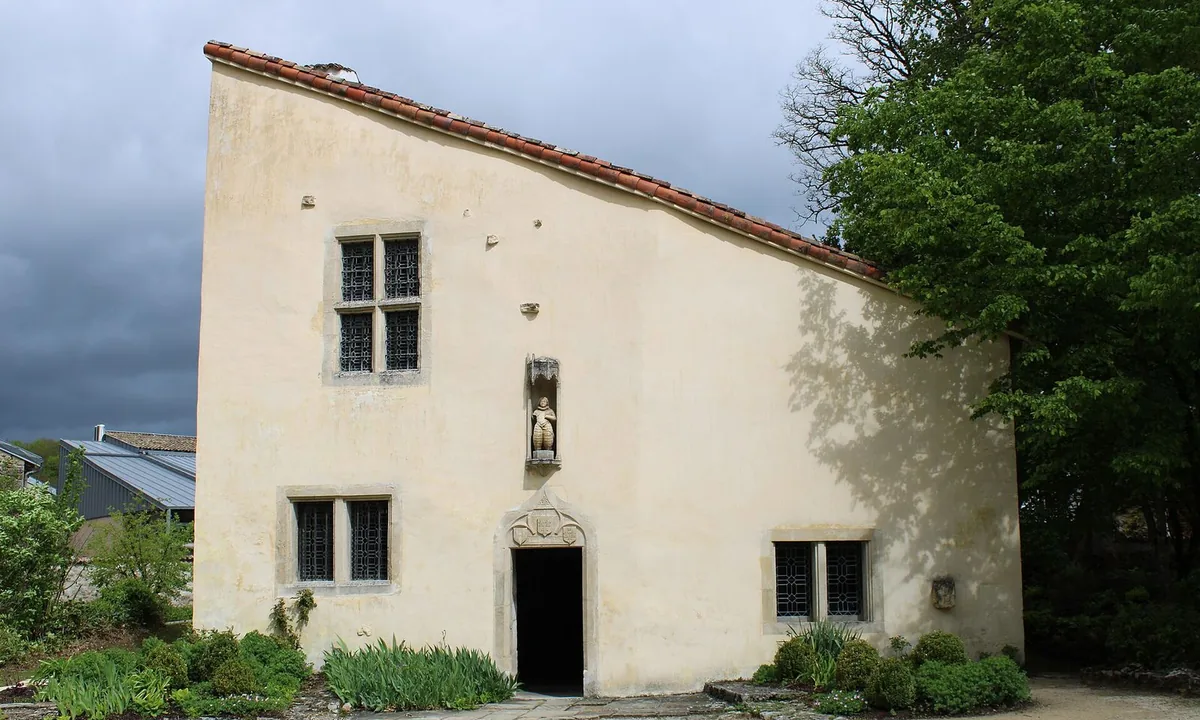 Old stone house with pale rendered walls, a long sloping tiled roof, small barred windows and a statue in a niche above the door, beside a gravel courtyard.