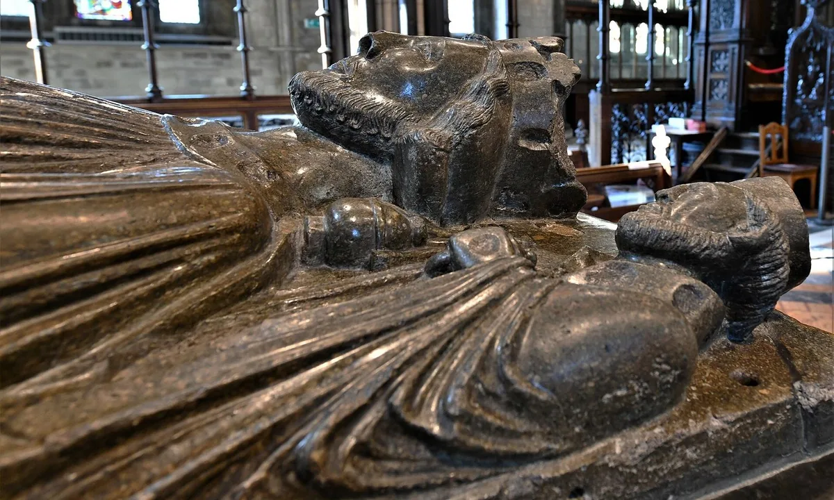 Carved stone figure of a crowned, bearded king lying on a tomb, holding a sword, with small figures by his head.