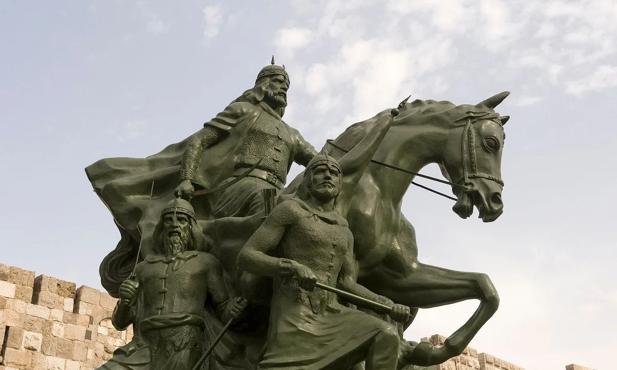 Dark bronze statue of a bearded rider on a rearing horse with two foot soldiers beside him, in front of the stone walls of a fortress.