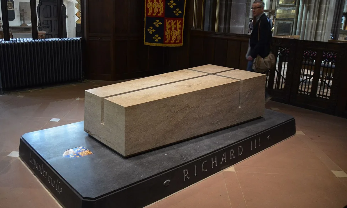 A pale stone tomb block cut with a deep cross, on a dark plinth carved RICHARD III, in a cathedral with a royal banner on the wall behind.