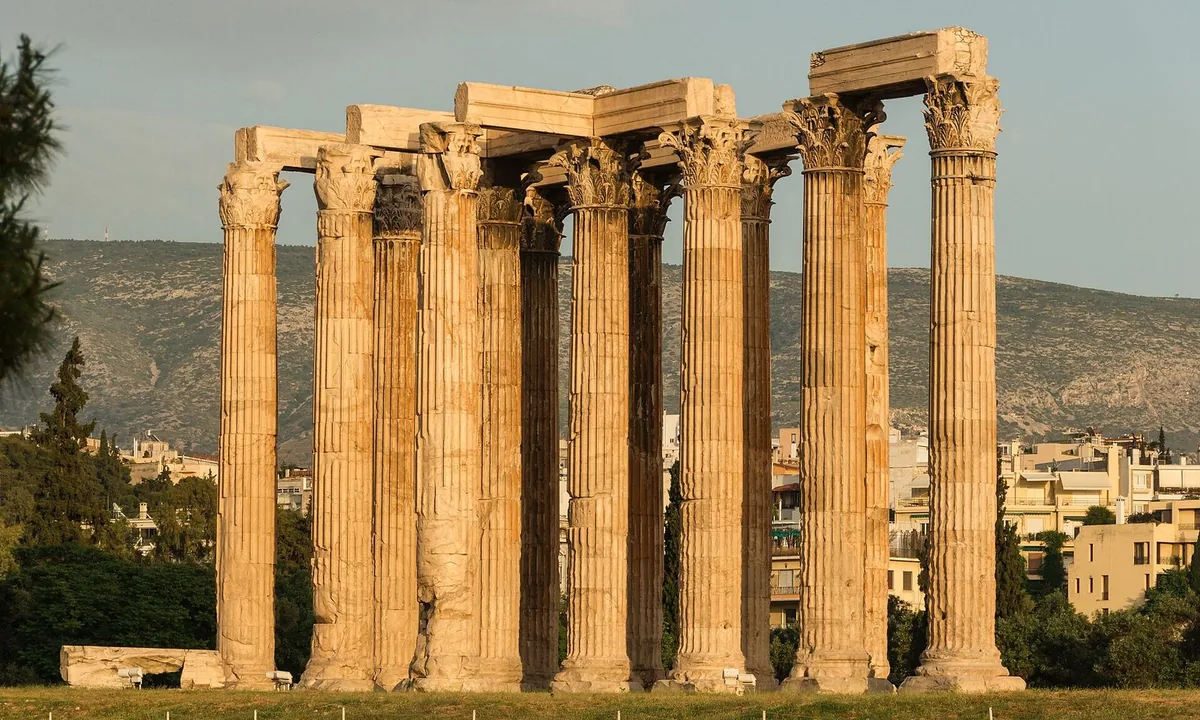 A group of tall fluted marble columns with ornate Corinthian tops, some still joined by stone beams, glowing in evening light against a hillside and modern buildings.