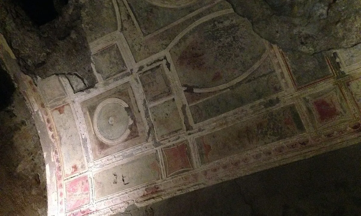 Curved ceiling of an underground Roman room painted with faded red and cream panels framing a central roundel, with darker unpainted plaster around it.