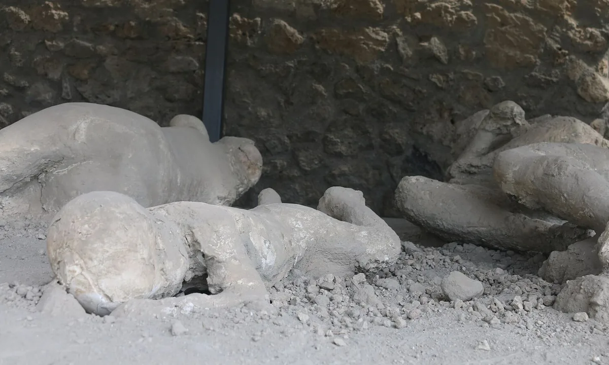 White plaster figures of several people lying curled on their sides on a bed of grey pumice, in front of a rough stone wall.
