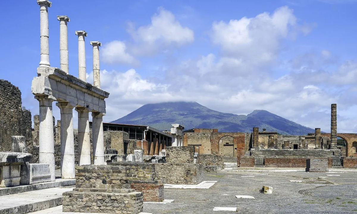 White columns and low stone walls on a flat paved plaza, with the broad cone of a mountain rising beyond under a blue sky with clouds.