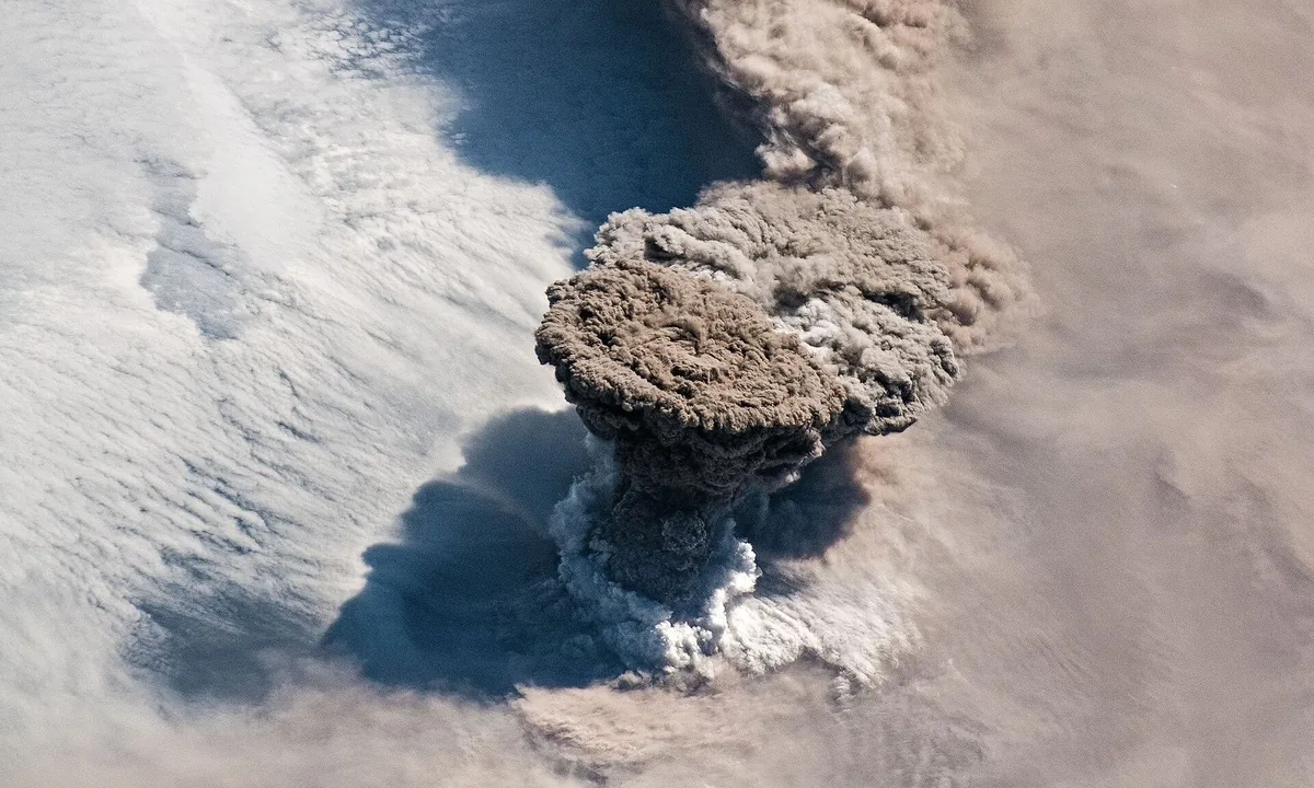 View from orbit of a column of brown-grey ash rising through white clouds and spreading out at the top like a mushroom.