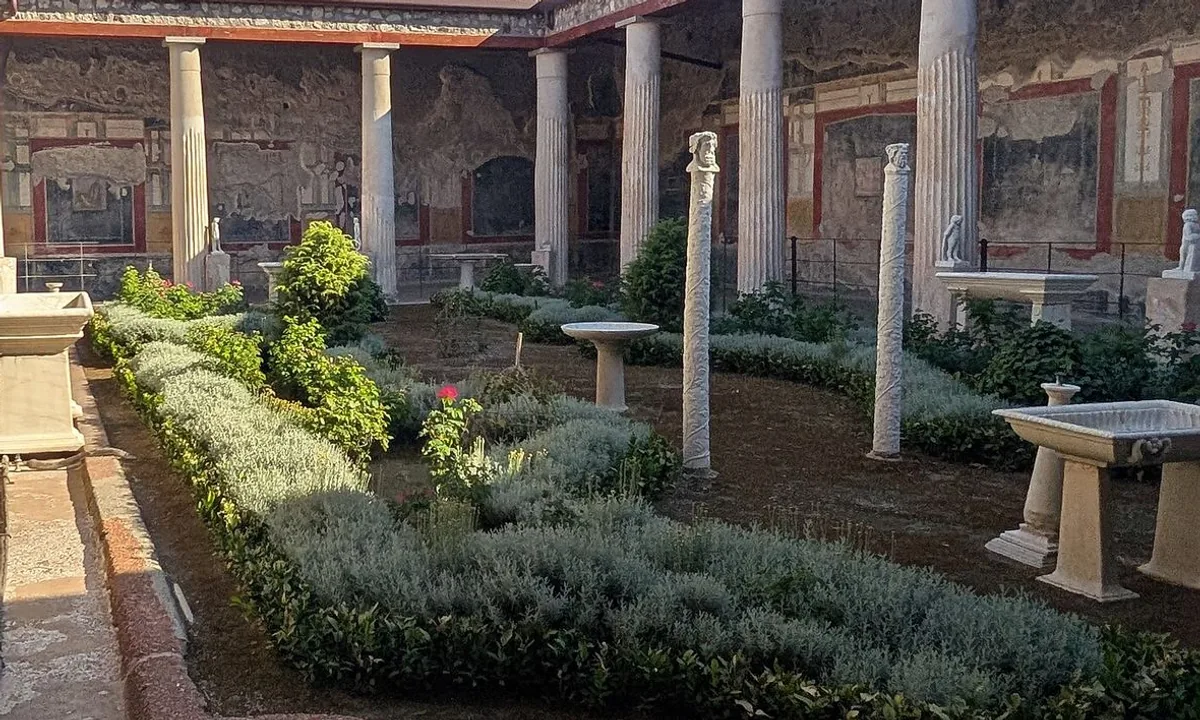 A garden courtyard of clipped hedges and small marble tables and fountains, surrounded by white columns, with red and black painted walls in the shade behind.