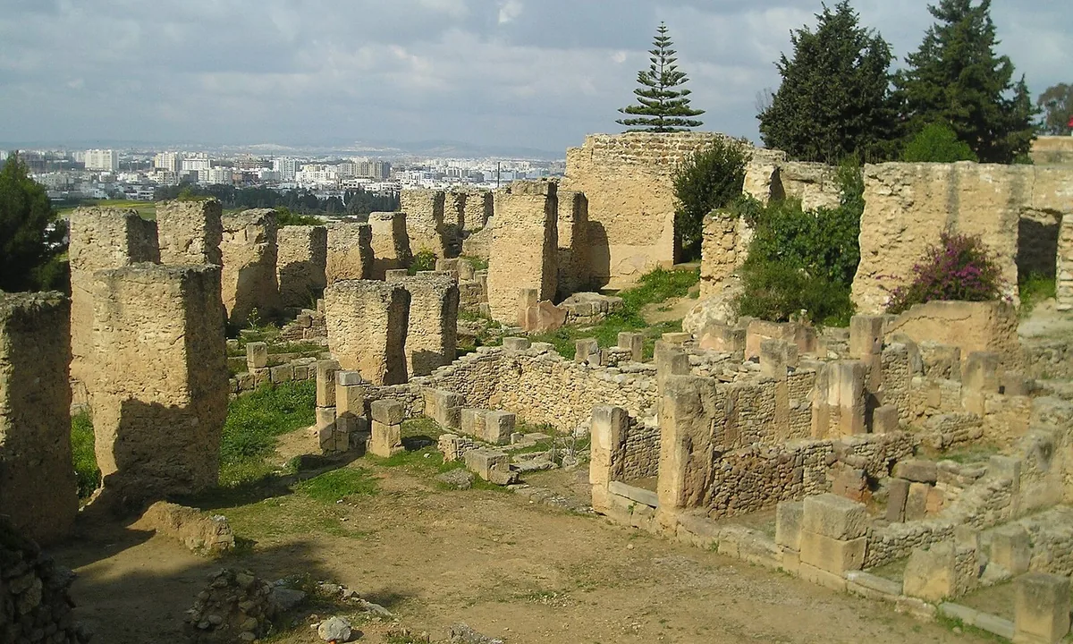 Excavated stone walls and floors of ancient houses on a hillside, with the sea and a modern town in the distance.