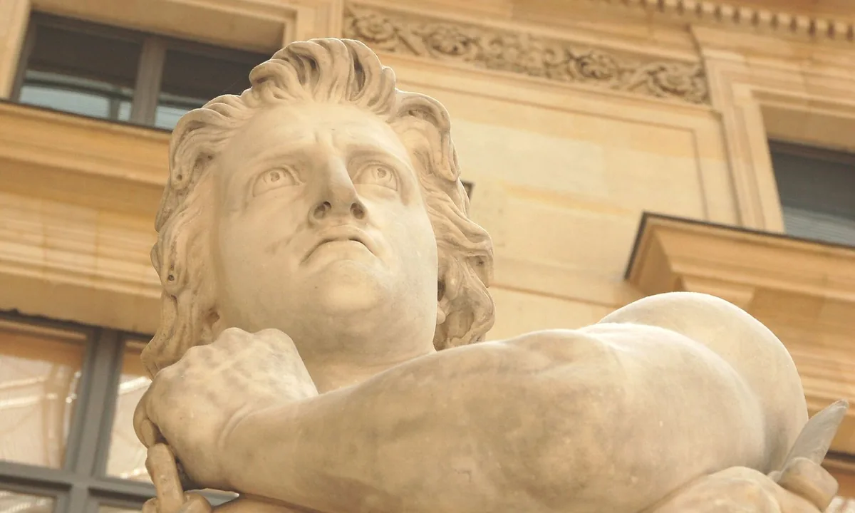 Marble statue of a man seen from below, with wavy hair and a defiant upward gaze, his muscular arms folded across his chest.