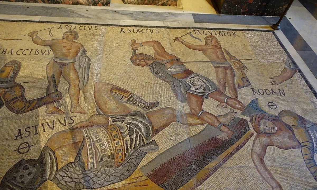Colourful floor mosaic of gladiators in armour fighting with swords and shields, some fallen, with their names written above them.