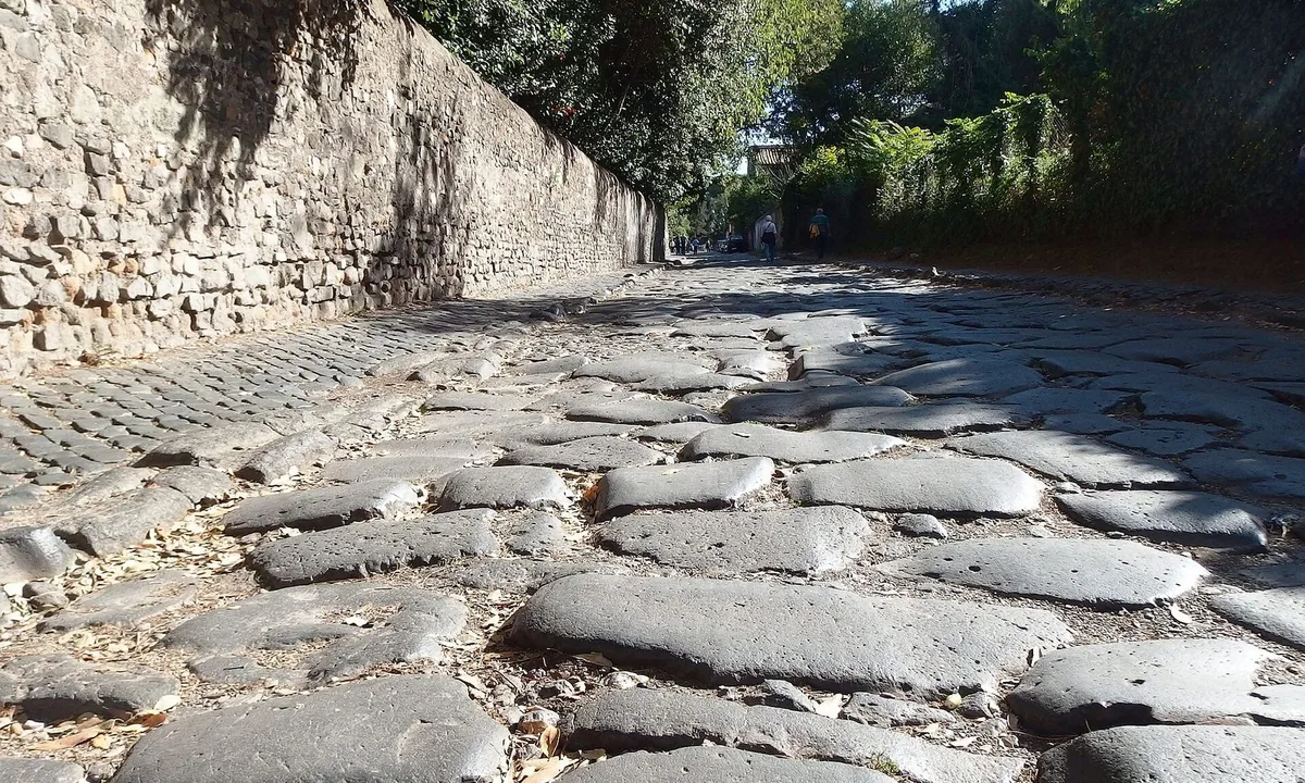 Ancient road of large, worn, dark paving stones running between a high stone wall and trees.