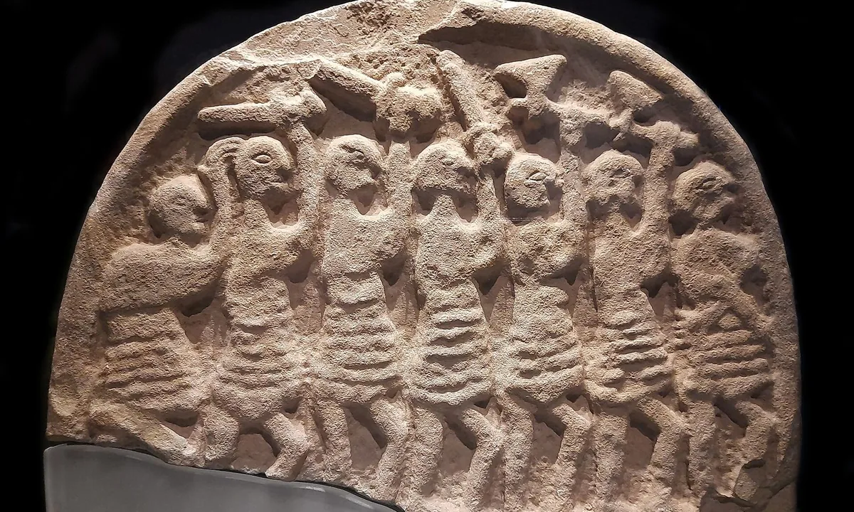 Weathered grey stone slab with a rounded top, carved with a row of figures raising weapons.