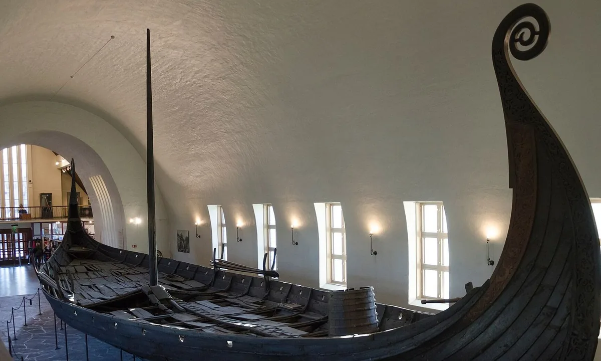 Long wooden ship with a high curling prow and stern, displayed in a white museum hall.