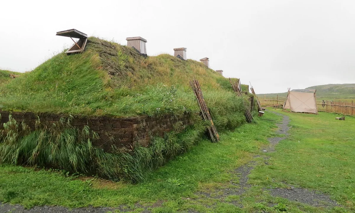 Low buildings with thick grass-covered turf walls and roofs on open coastal grassland under a grey sky.