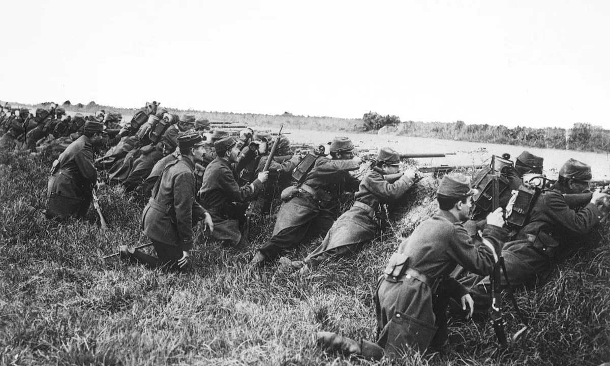 Black-and-white photo of a line of French soldiers in kepis and greatcoats kneeling and lying behind a grassy bank, aiming rifles.