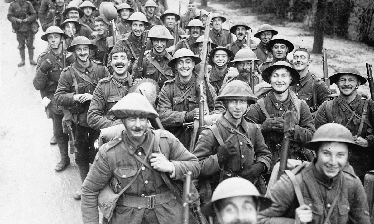 Black-and-white photo of a column of smiling British soldiers in steel helmets with rifles, marching along a country road towards the camera.